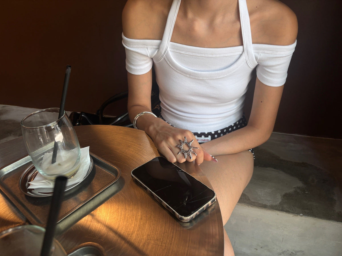 Mujer con top blanco y anillo grande de acero en forma de estrella, sentada en cafetería.