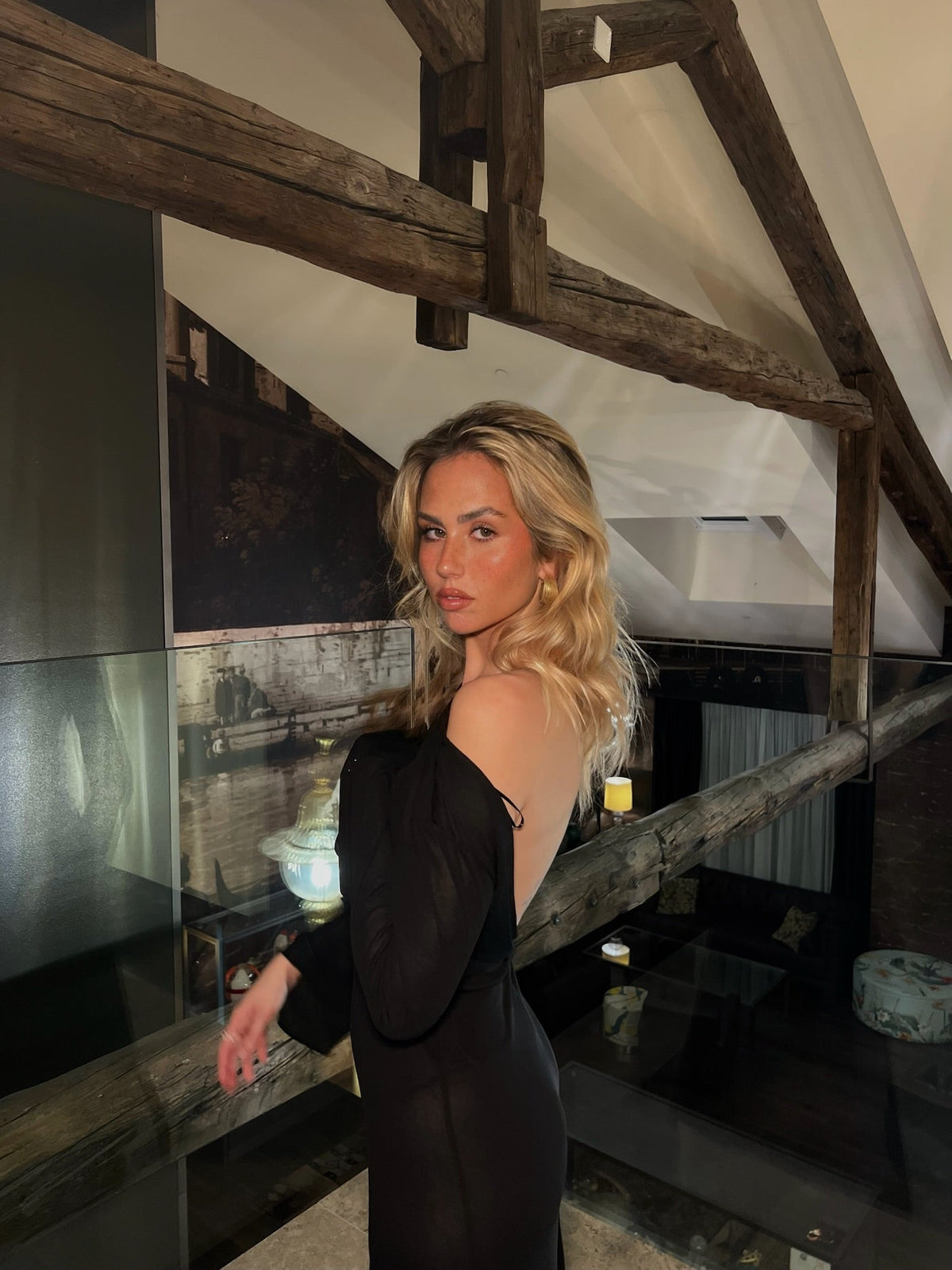 Mujer rubia con vestido negro elegante en interior moderno con vigas de madera visibles.