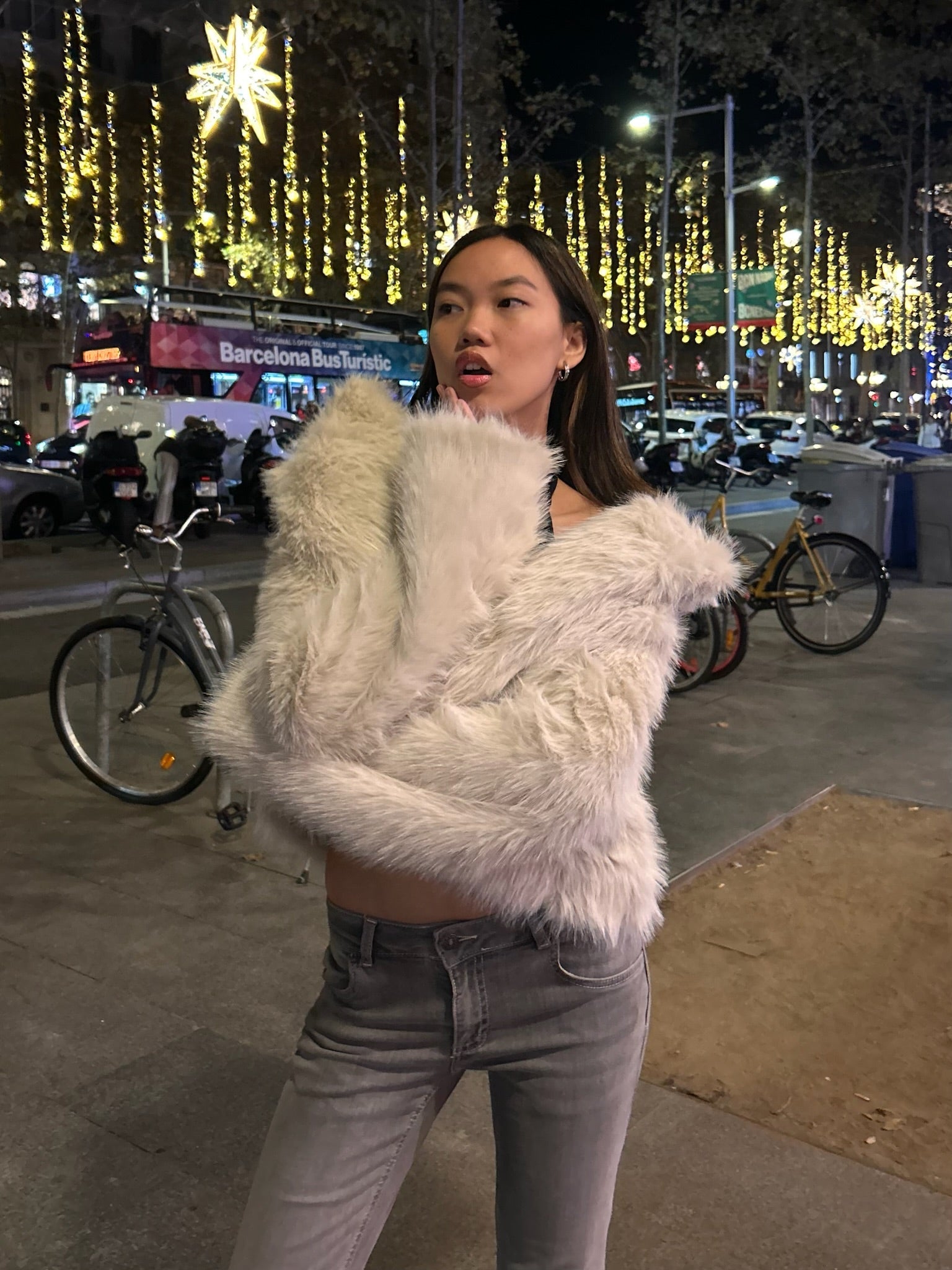 Mujer con abrigo de peluche blanco y jeans grises en ciudad iluminada de noche, estilo moderno