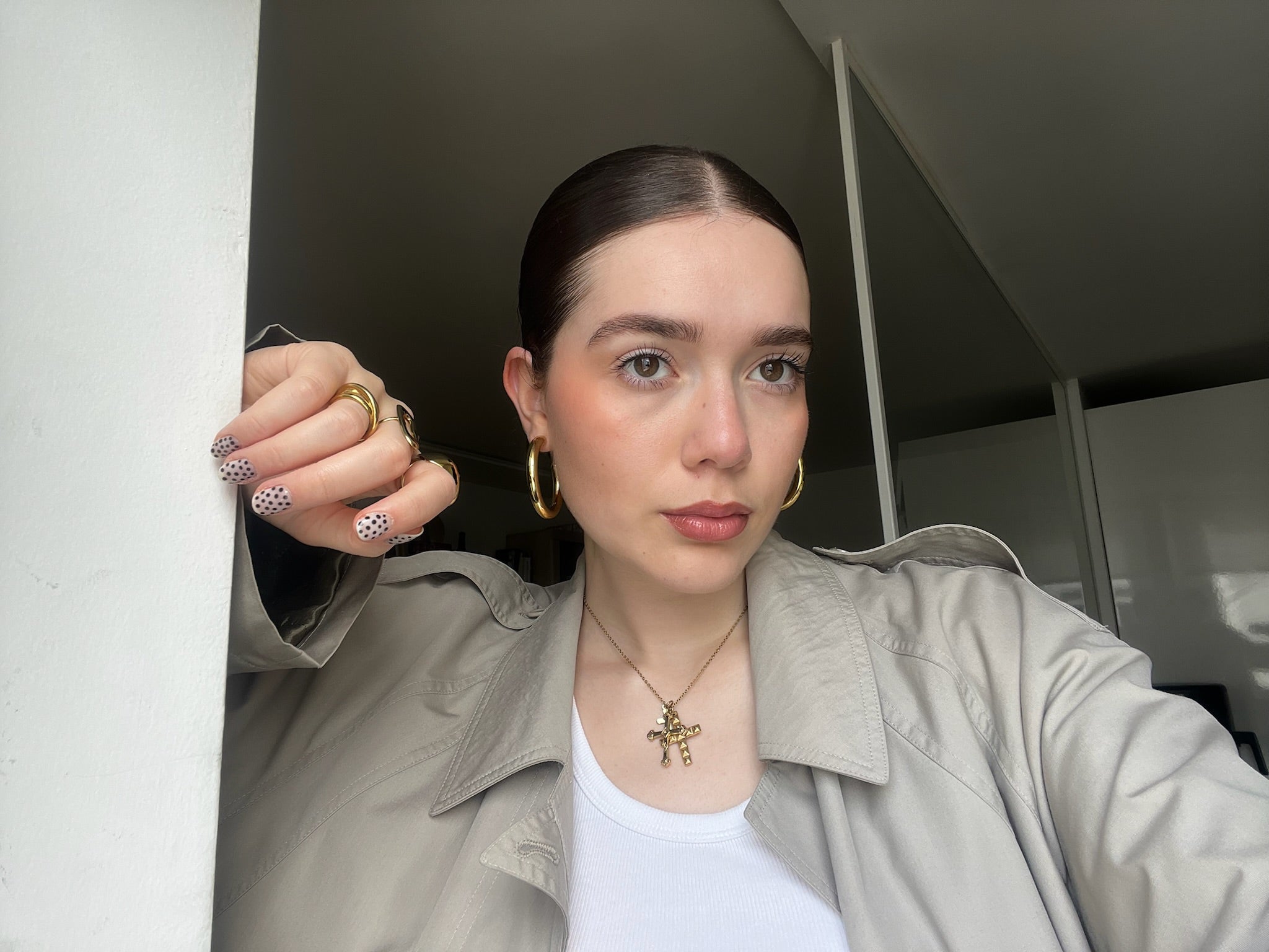Mujer con joyas de acero dorado, anillos y aretes hipoalergénicos, manicura moderna y gabardina beige.