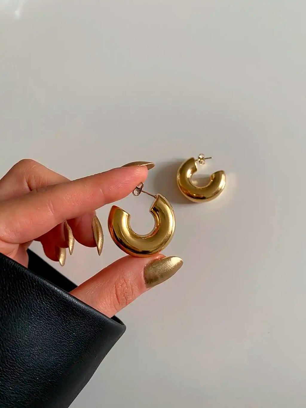 Pendientes Aro Big acero inoxidable dorado, lo muestra una mano de mujer con las uñas doradas. Su diseño es semi-circular con un corte abierto que aporta un toque contemporáneo y minimalista. - DELACABRA