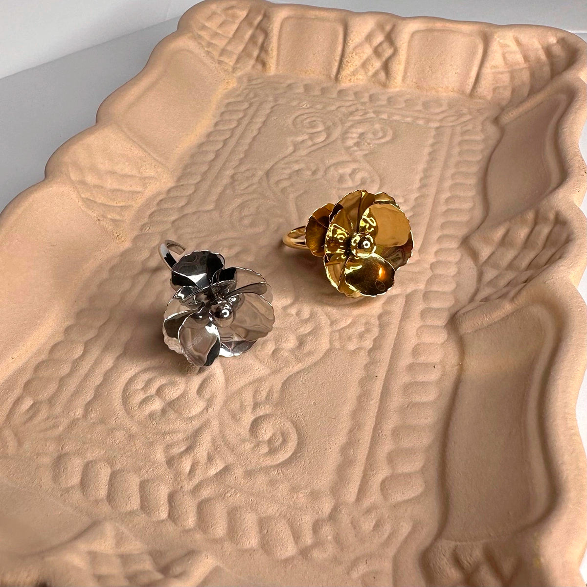 Anillos de acero inoxidable en forma de flor, uno dorado y otro plateado, sobre bandeja decorativa.