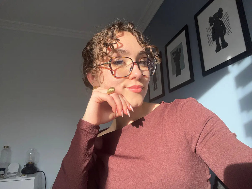 Mujer usando el anillo DROP DELACABRA en acabado dorado, diseño ajustable con forma de gota, fotografiada con luz natural junto a ilustraciones decorativas