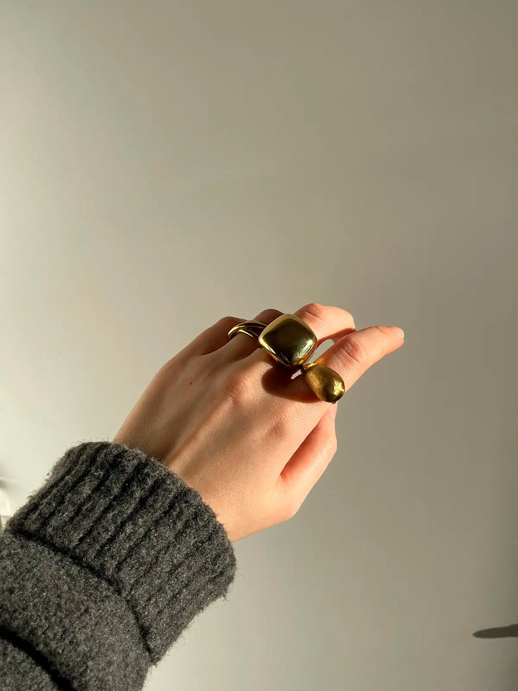 Mano con varios anillos dorados ajustables de acero inoxidable puestos, fotografiada con luz natural y fondo neutro, look invernal con jersey gris