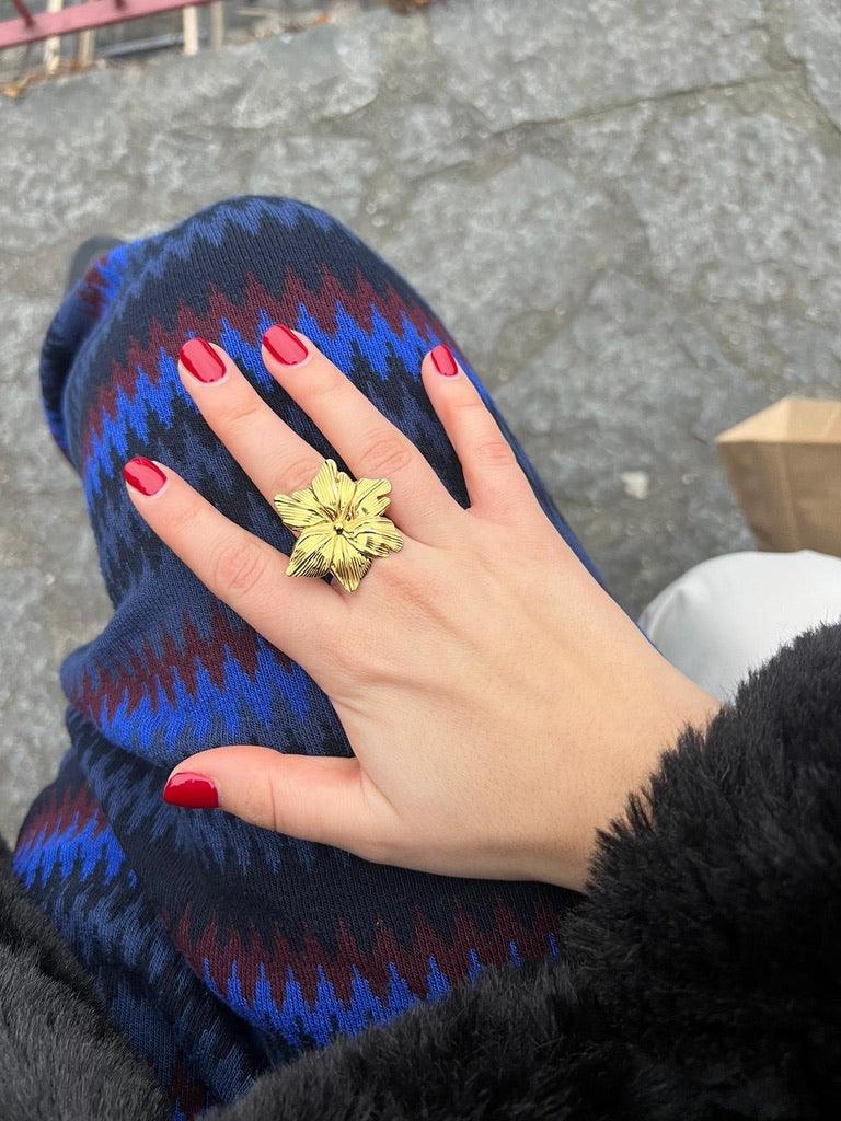 Anillo Hawai marca delacabra, tono dorado en forma de flor tropical, acero inoxidable ajustable, anillo en mano de una modelo con uñas rojas