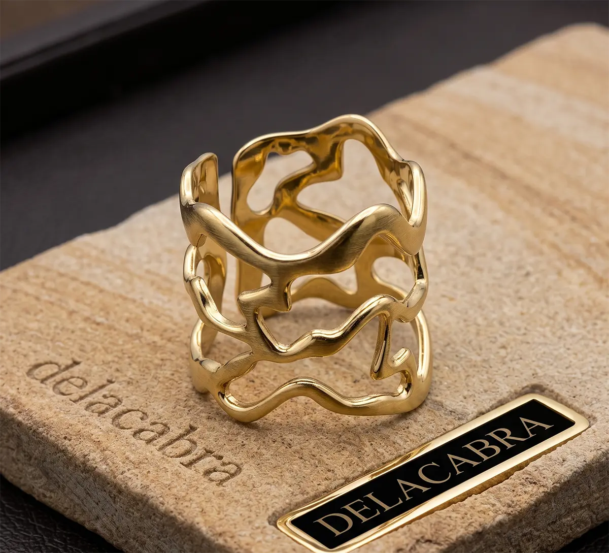 Anillo Tango acero inoxidable dorado acabado brilante, sobre una superficie de roca labrada con el logo de delacabra y fondo oscuro elegante. Delacabra