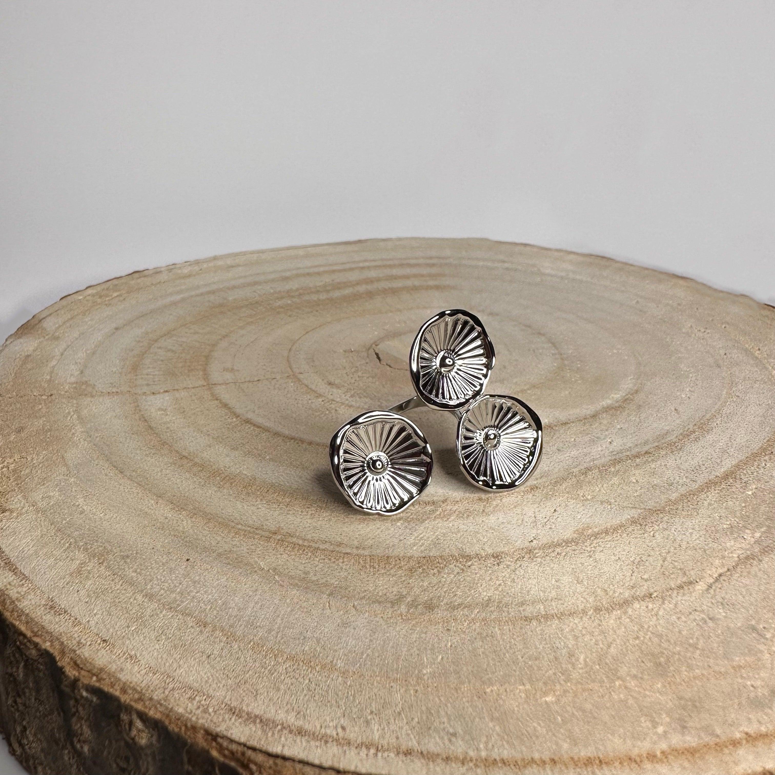 Anillo ajustable de acero inoxidable plateado con diseño de tres flores sobre fondo de madera