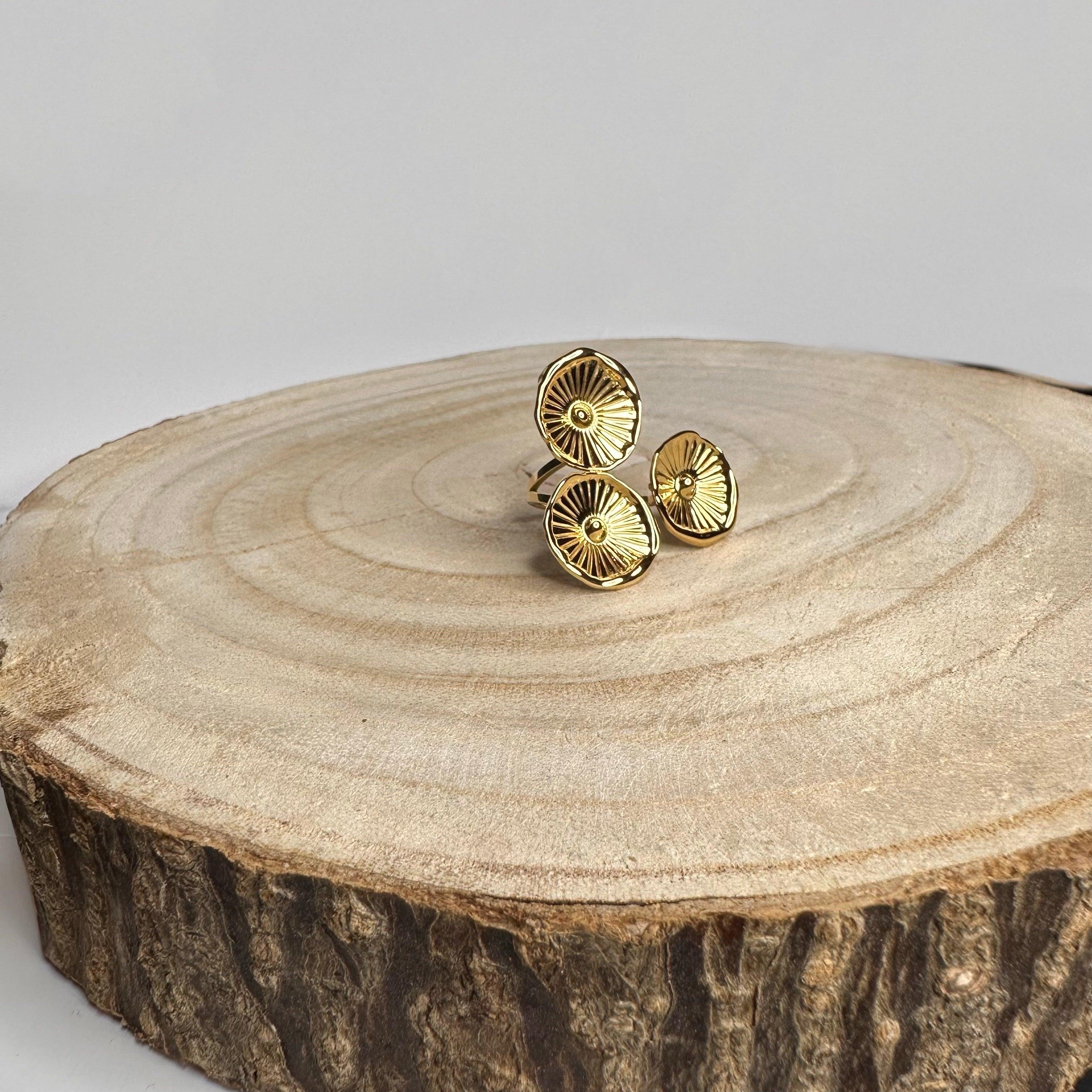 Anillo dorado ajustable de acero inoxidable con diseño floral, sobre base de madera.