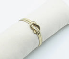 Brazalete Tangled acero inoxidable dorado entrelazado simulando un nudo llano, acabado suave y brillante en expositor de tela - DELACABRA