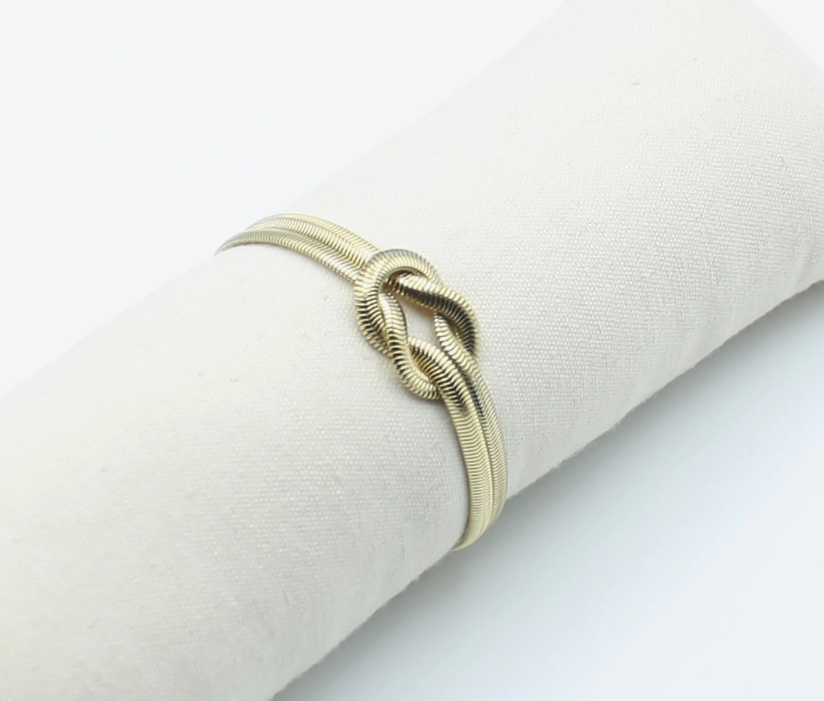 Brazalete Tangled acero inoxidable dorado entrelazado simulando un nudo llano, acabado suave y brillante en expositor de tela - DELACABRA