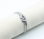 Brazalete Tangled acero inoxidable plateado entrelazado simulando un nudo llano en un expositor de tela, acabado suave y brillante - DELACABRA