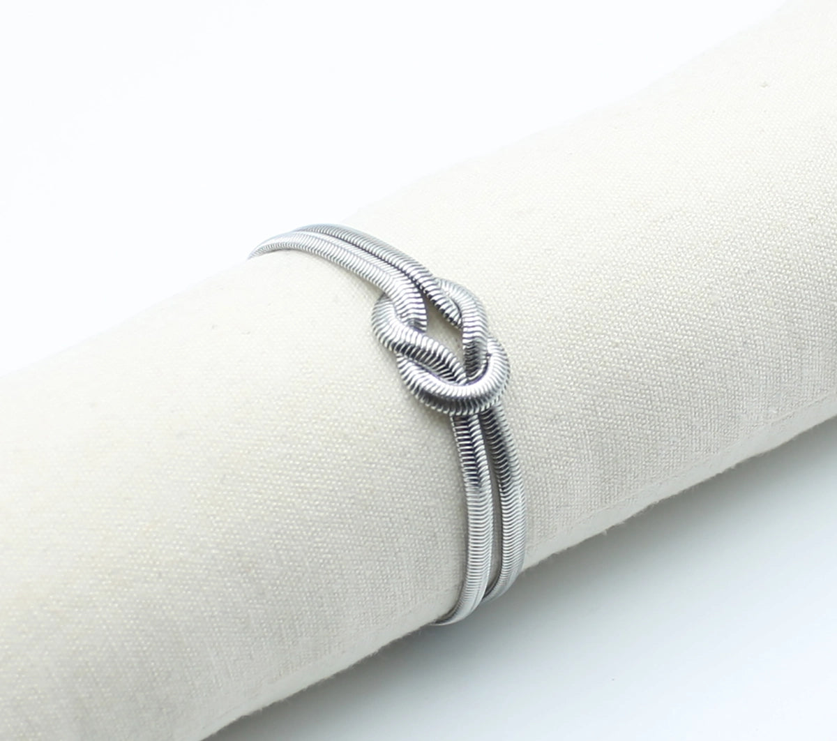Brazalete Tangled acero inoxidable plateado entrelazado simulando un nudo llano en un expositor de tela, acabado suave y brillante - DELACABRA