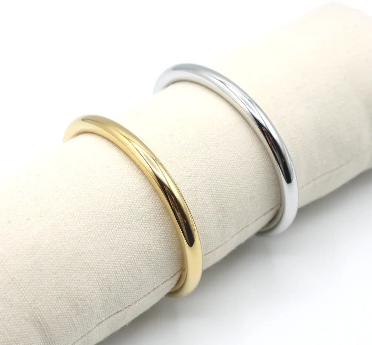 Pareja de brazaletes Lia en versiones dorada y plateada sobre exhibidor textil, mostrando su diseño tubular minimalista y estructura ajustable de acero inoxidable.