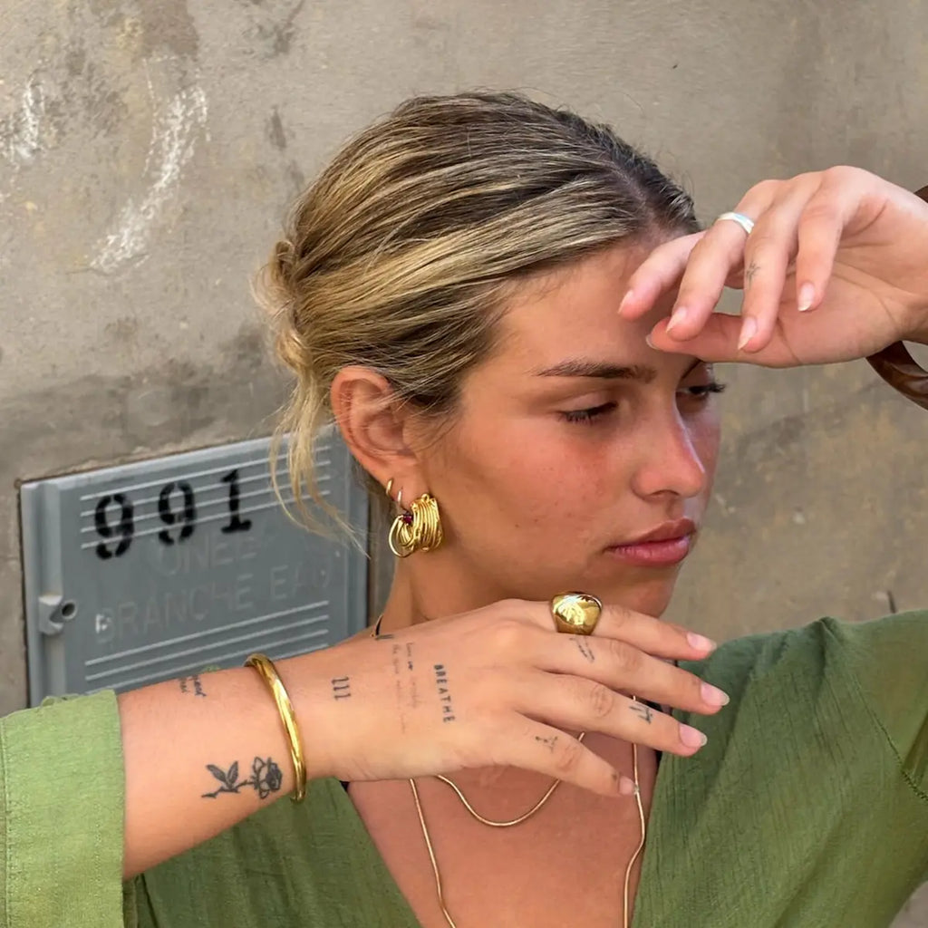 Look con joyas doradas DELACABRA en acero inoxidable hipoalergénico y resistente al agua: pendientes de aro múltiples, anillo de sello en relieve, pulsera rígida minimalista y collar tipo cadena serpiente.