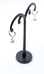 Pendientes Aro Nácar plateados con perla blanca colgante, acero inoxidable hipoalergénico y resistente al agua, vistos en expositor negro.