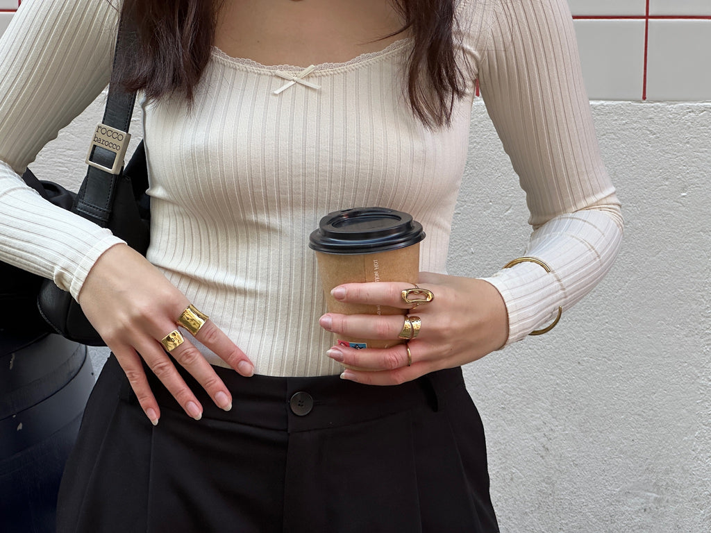Mujer con varias joyas doradas de acero inoxidable de la marca DELACABRA, incluyendo anillos, brazalete y pendientes ajustables, hipoalergénicos y resistentes al agua, en un look urbano con café en mano