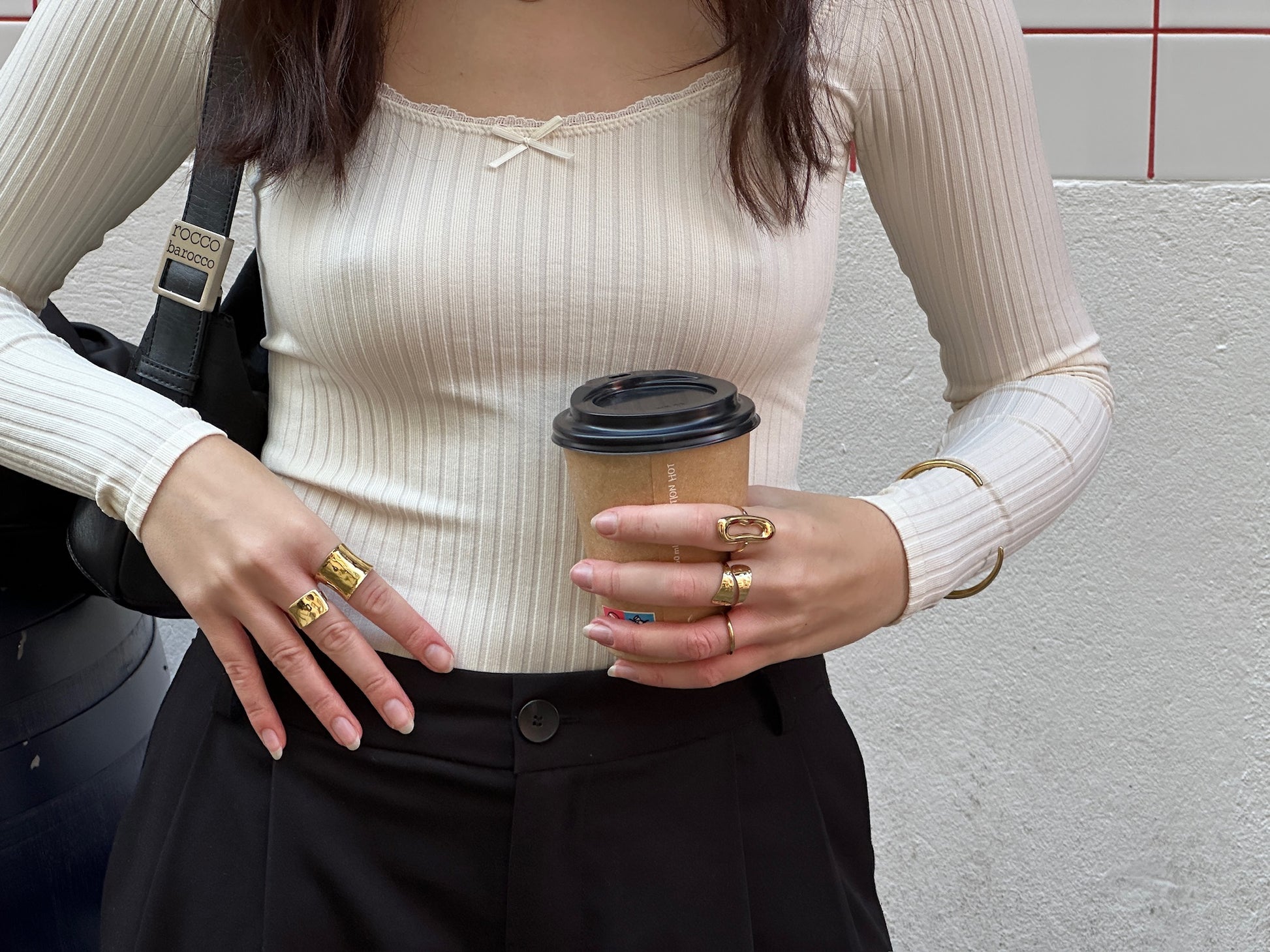 Mujer con varias joyas doradas de acero inoxidable de la marca DELACABRA, incluyendo anillos, brazalete y pendientes ajustables, hipoalergénicos y resistentes al agua, en un look urbano con café en mano