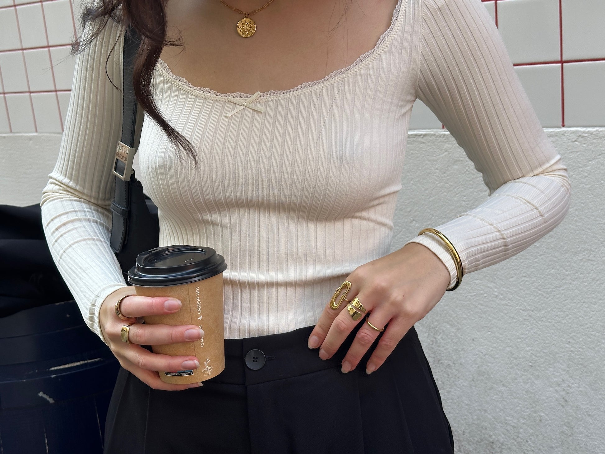 Mujer con joyas doradas de acero inoxidable de la marca DELACABRA, incluyendo anillos, pulsera y collar ajustables, hipoalergénicos y resistentes al agua, sujetando un café en la calle