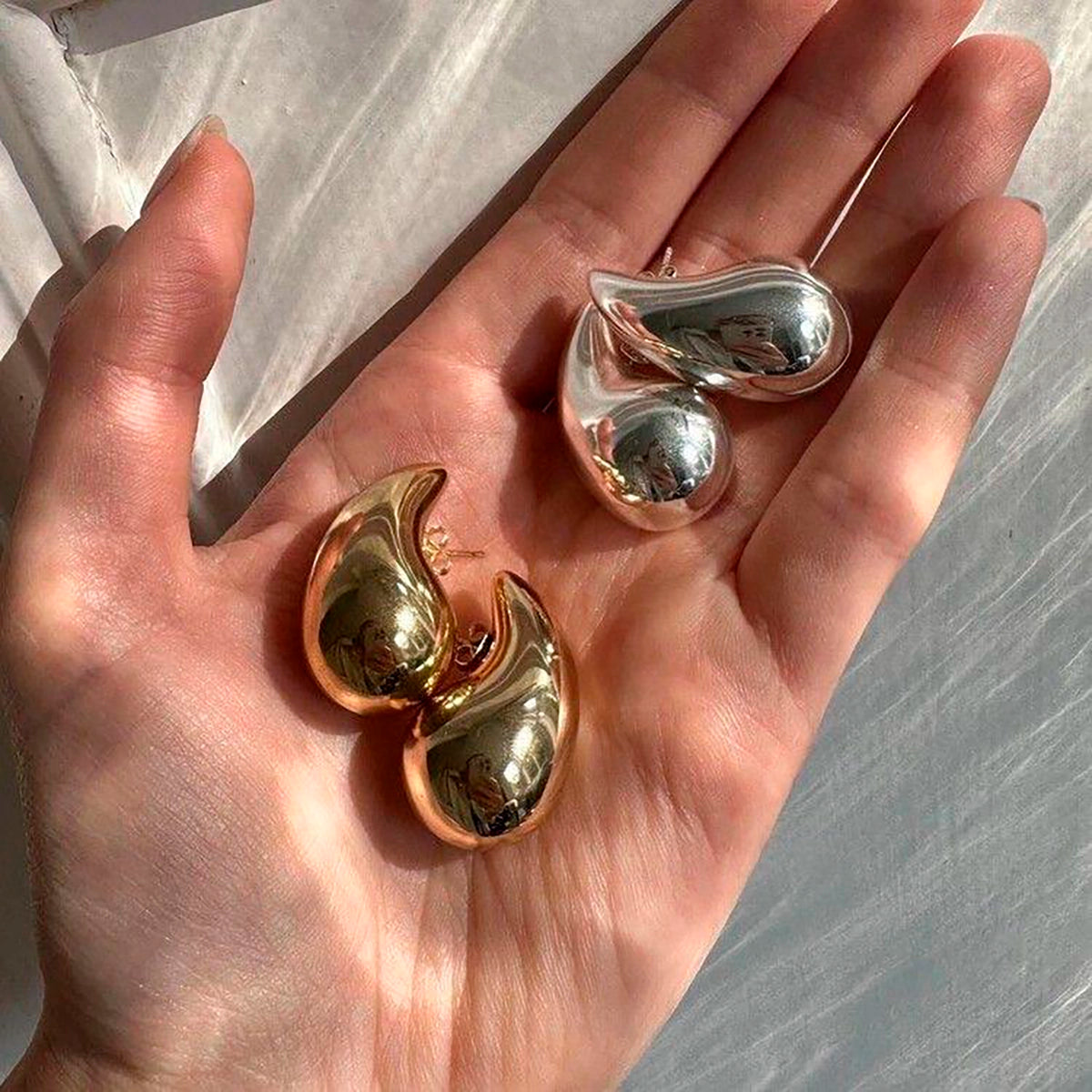 Pendientes de gota grandes dorados y plateados de acero, sostenidos en una mano femenina.
