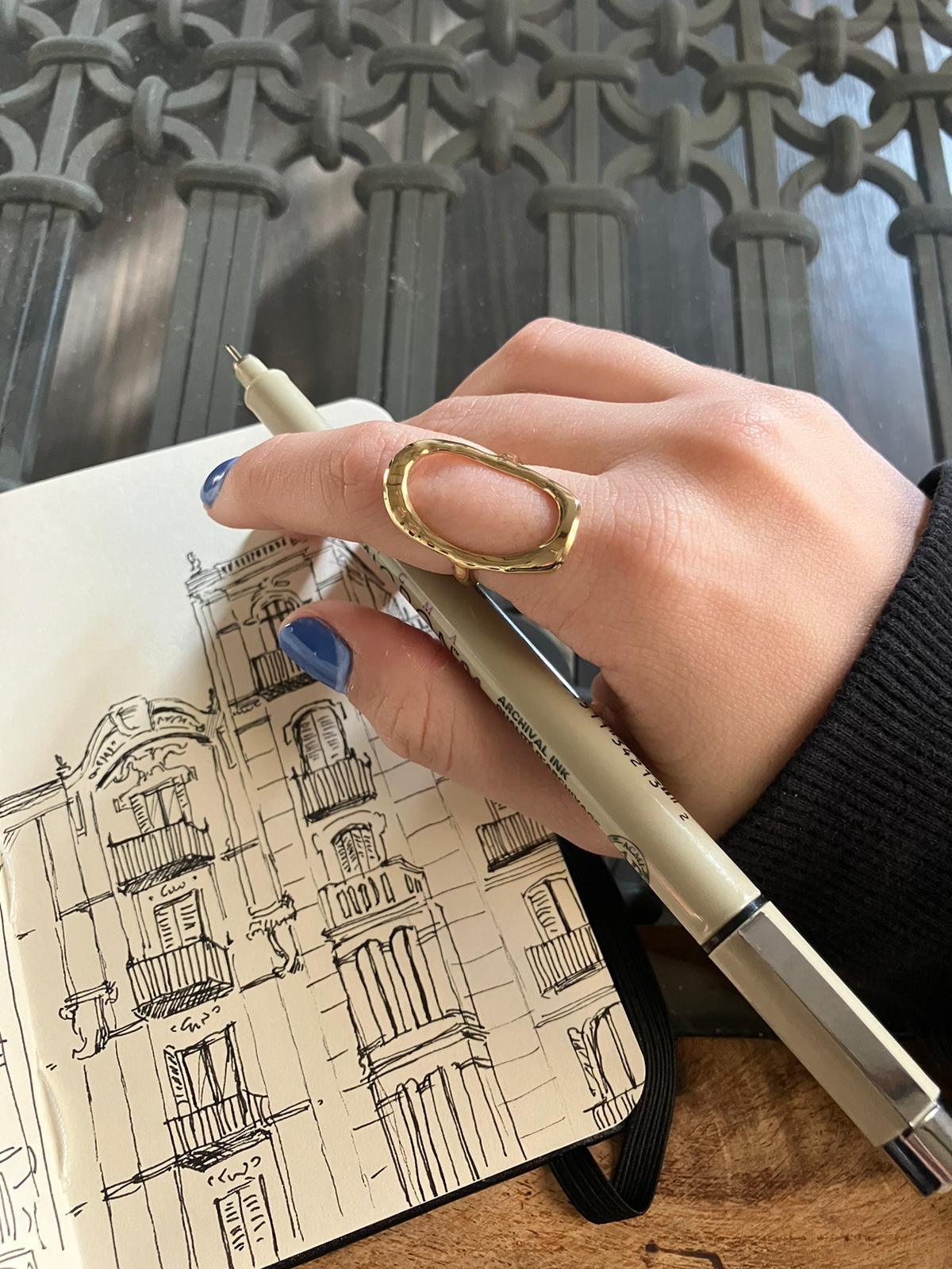 Anillo dorado ajustable de acero inoxidable de la marca DELACABRA puesto en la mano de una mujer mientras dibuja arquitectura