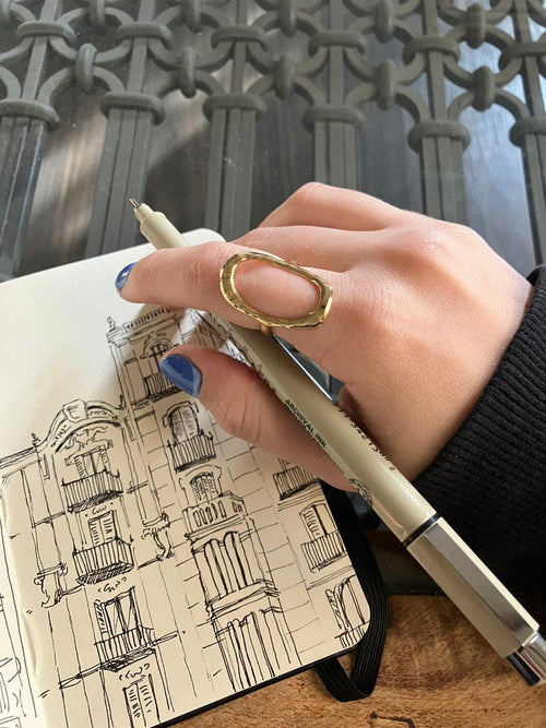 Anillo dorado grande en mano femenina dibujando edificio en libreta, joya contemporánea